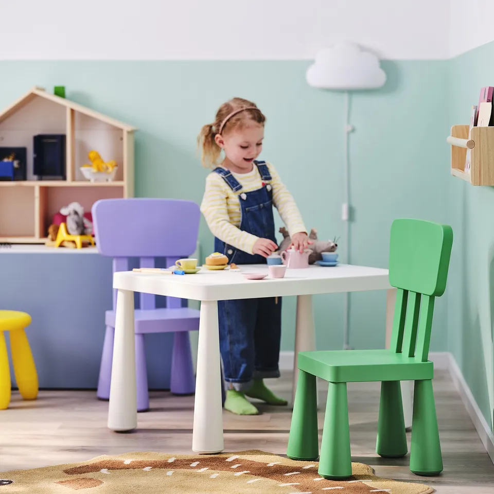 Child in overalls playing with toy teacups at a white table on a rug, surrounded by colourful chairs for indoor/outdoor tea parties.