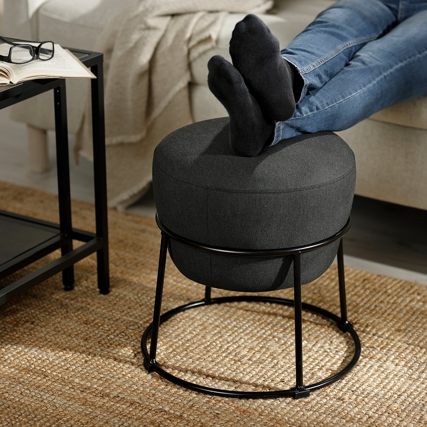 Person resting feet on round black stool; stool has metal frame, cushioned seat.