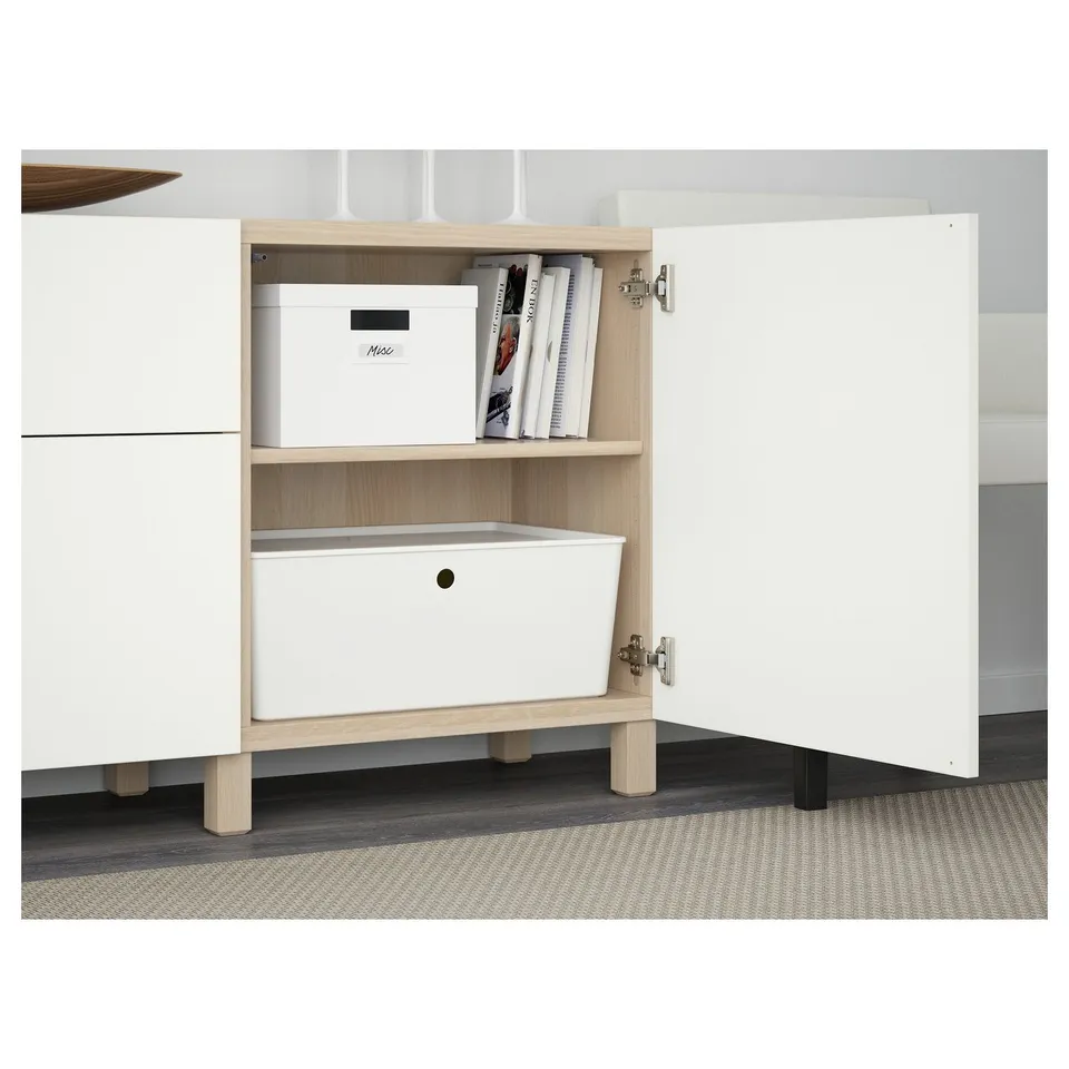 White cabinet with light wood trim, two open sections. Top shelf holds white box and books. Bottom has a large white bin on small legs.