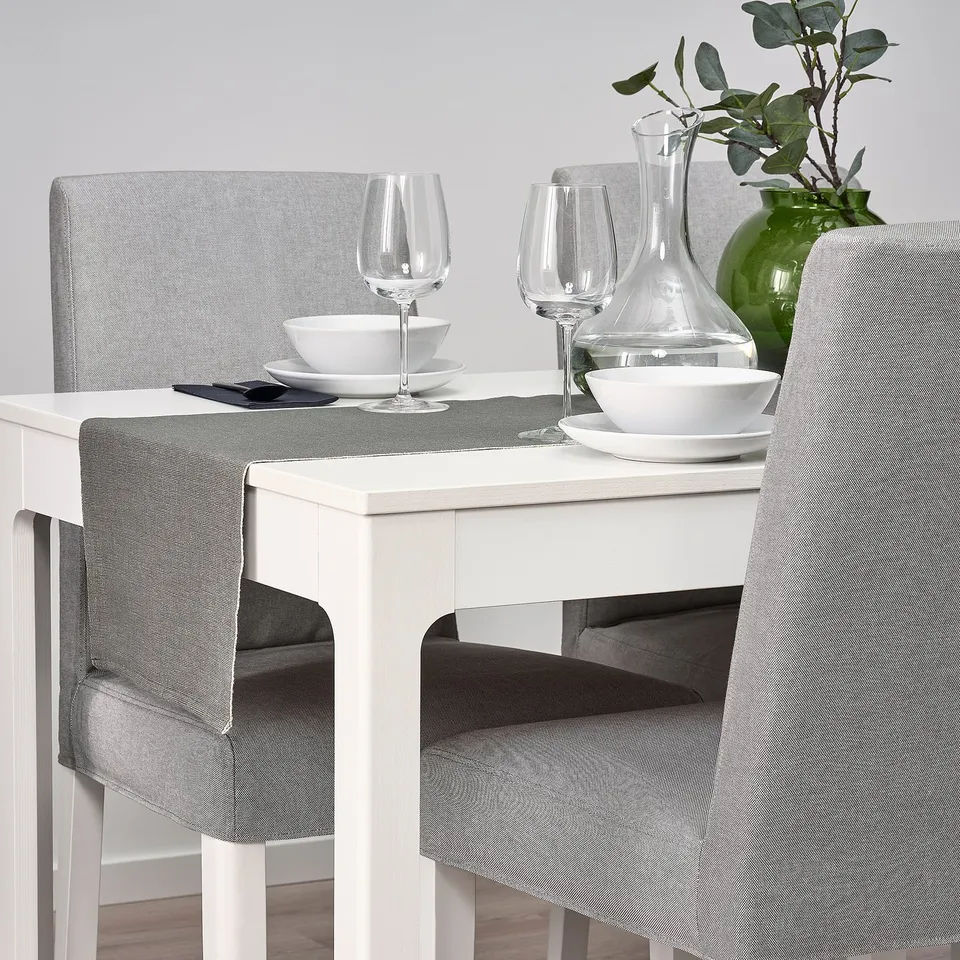 Table set with white plates, glasses, and carafes atop a grey table runner on a white table, framed by grey and white upholstered chairs.