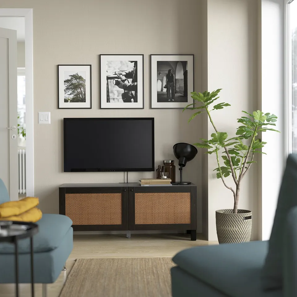 A modern living room with a black BESTÅ tv bench & wicker, decorated with photos, books, a lamp, a plant, and a yellow blanket on a blue chair.