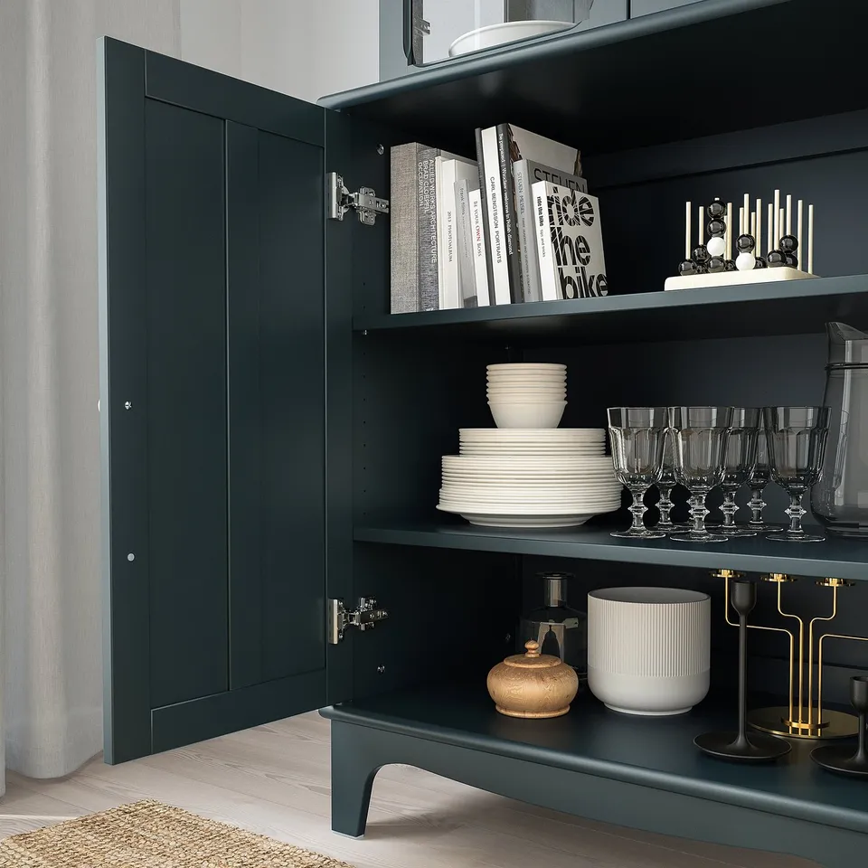 Dark wooden cabinet with glass doors showcasing stacked plates, glasses, decorative vases, and books. Soft-close hinges visible.