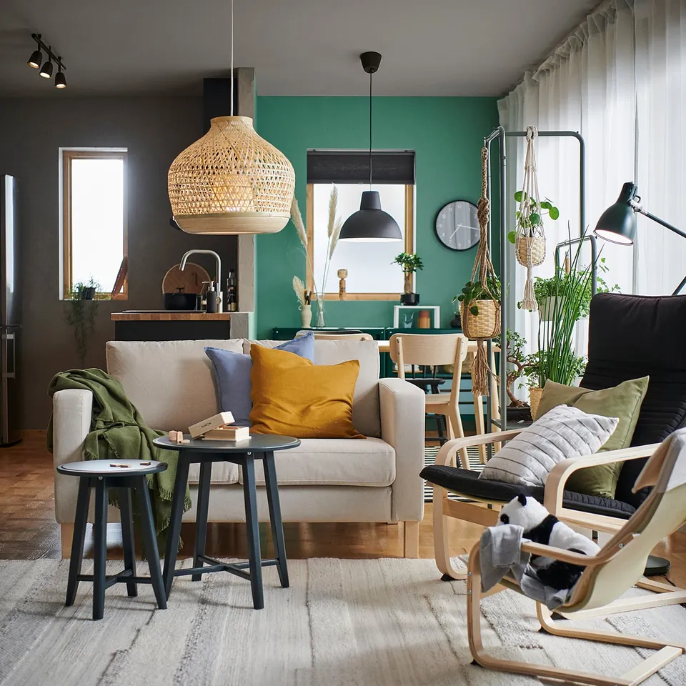 Modern living room with green wall, grey sofa, colourful pillows, wooden floor, plants, and lamps.