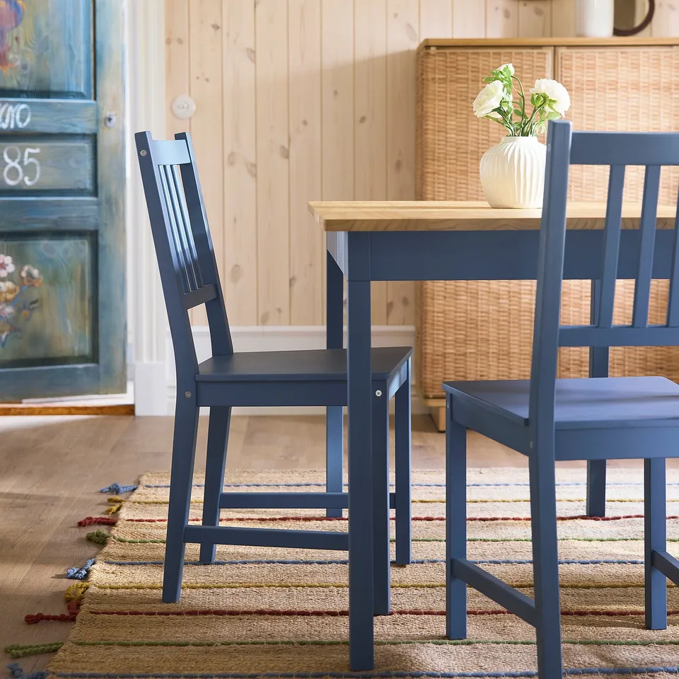 Cosy dining area with 2 dark blue wooden PINNTORP chairs, rectangular table, floral vase, woven rug, beside wooden door.