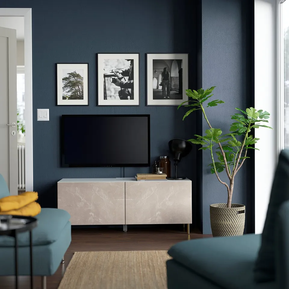 Modern living room with BESTÅ tv bench, flat screen, and plants. Blue chairs, natural rug, framed photos.