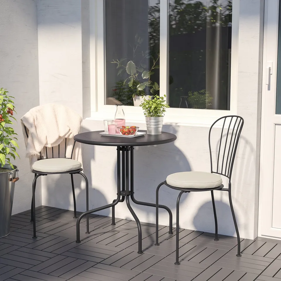 Small outdoor table with two matching chairs. Black metal frame, white cushions. Situated near a window with potted plants.