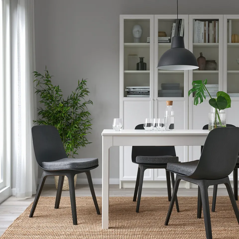 Small grey chair pads sit on black modern chairs around a white dining table, contrasting with larger green plants and open shelves behind.