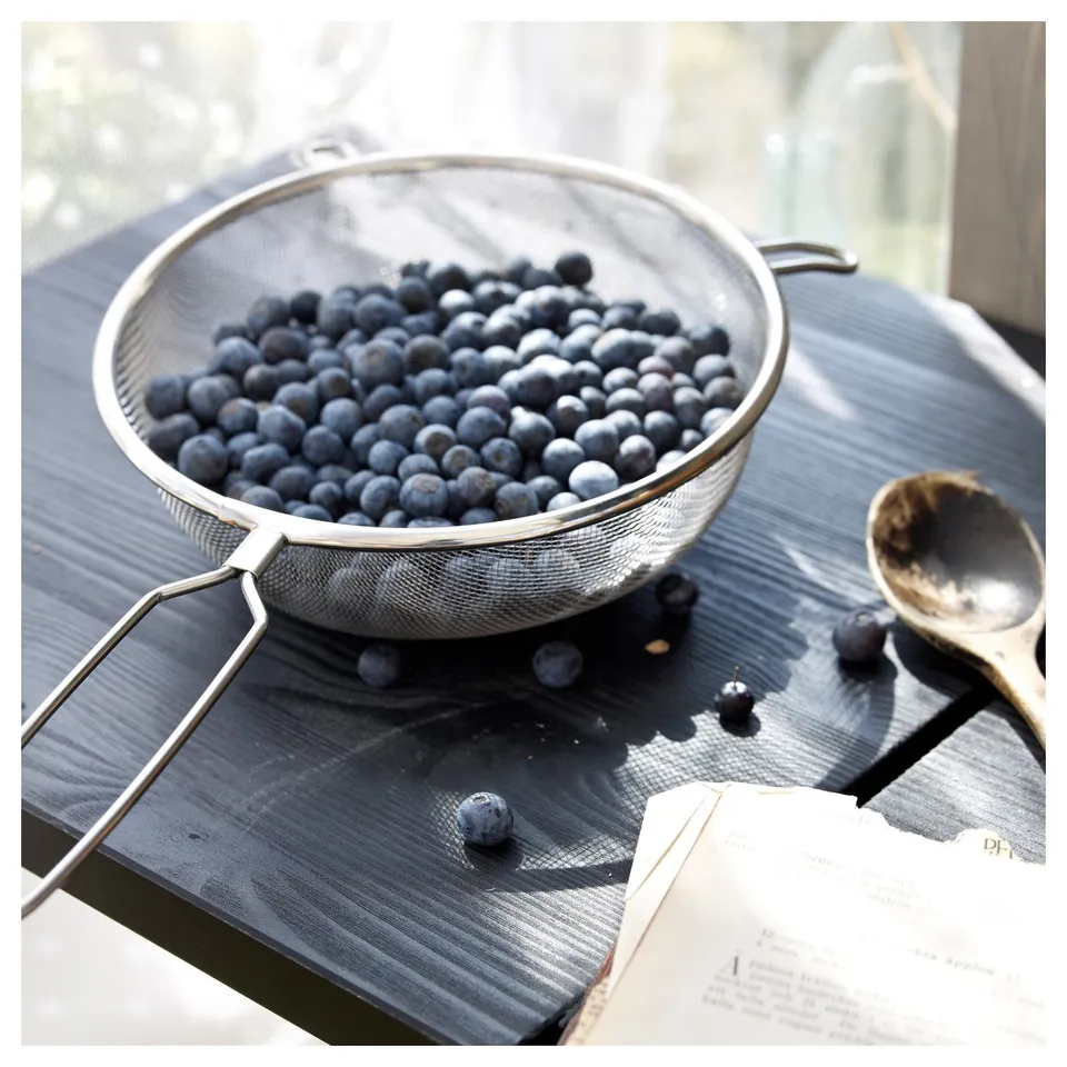 A stainless steel strainer full of blueberries on a wooden table with a spoon and an open book.