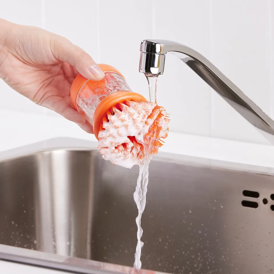 A hand holds an orange brush under a tap, dispensing dish soap onto dishes, showcasing its dual functionality of brushing and soap dispensing.