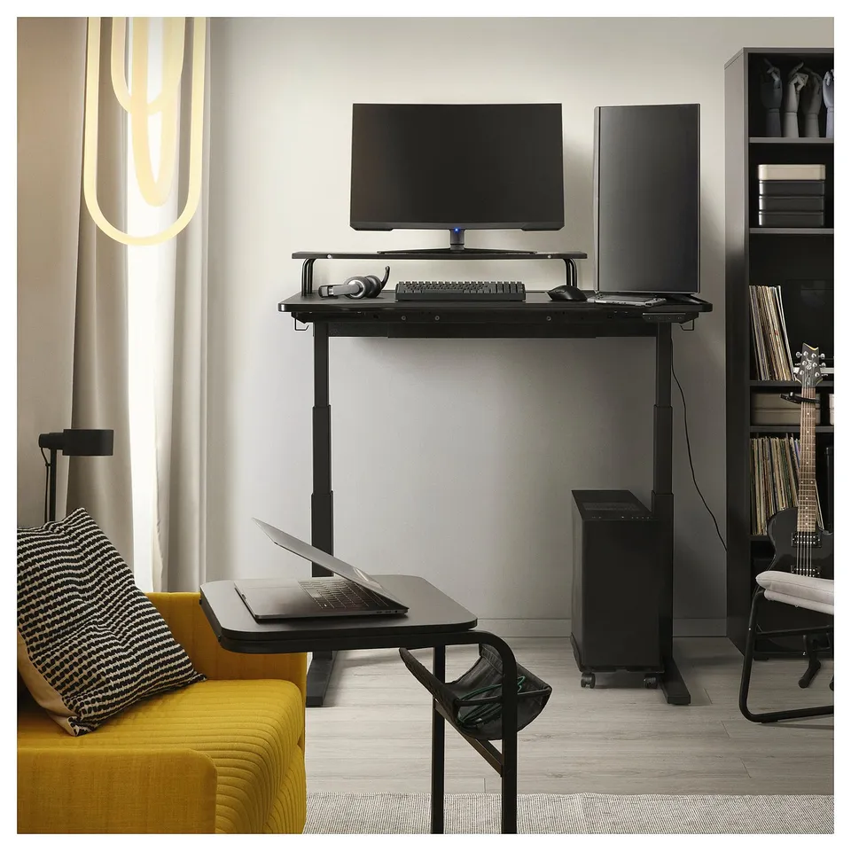 Modern gaming setup with black adjustable desk, monitor, laptop, and yellow chair.