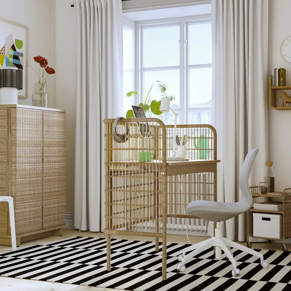 Home office with FRYKSÅS desk, wicker rack, striped rug, laptop, books, vases, curtains, and window.