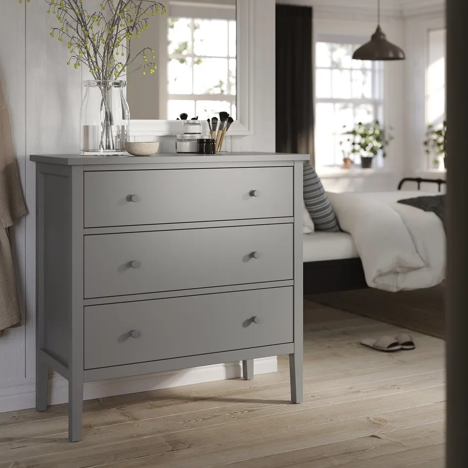 Grey GULLABERG chest with 3 drawers, knobs, legs, vase, makeup, near bed, window.