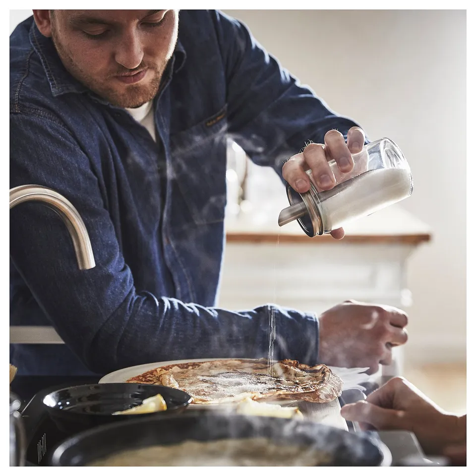 Person shakes sugar from CITRONHAJ, clear container, stainless lid, onto food.