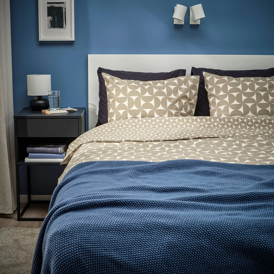 Modern bedroom with blue wall, beige patterned bedding, black bedside table with lamp and books, framed picture on wall, white headboard, wall lights.