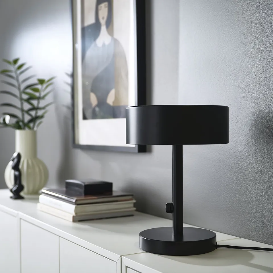NYMÅNE lamp on table with books and plant. Frosted glass diffuses warm light.