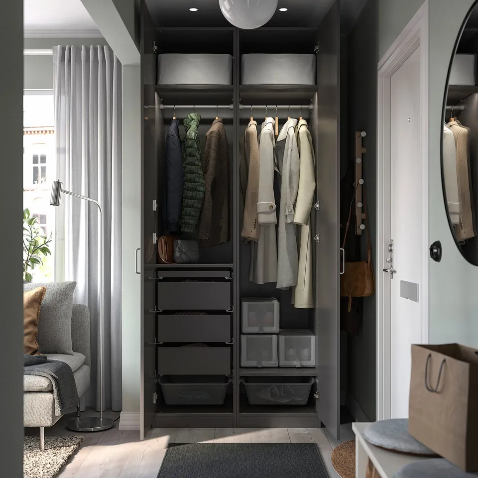 Modern bedroom with PAX wardrobe in dark finish, holding clothes and accessories, adjacent to a grey sofa and a large round mirror.