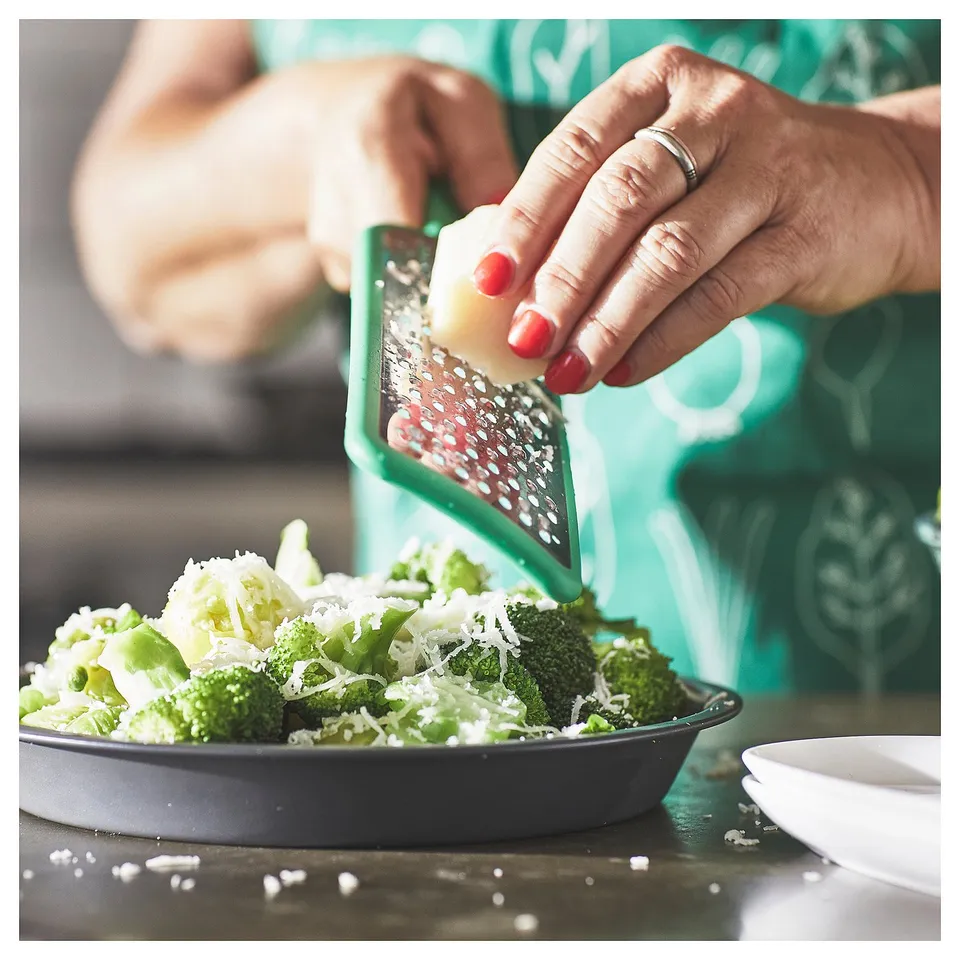 A person is grating cheese over a bowl of broccoli using a green UPPFYLLD grater. The grater has a green handle and a stainless steel grating surface.