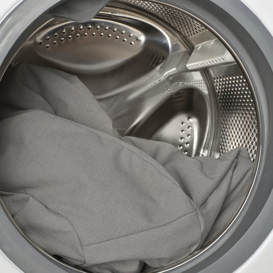 Washing machine drum with open lid, containing crumpled grey fabric.