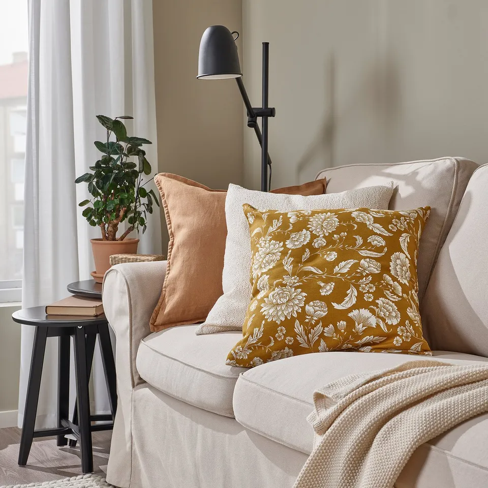 Cosy living room with light sofa & mustard yellow floral cushions. Black side table with plant & books, adjustable lamp nearby.