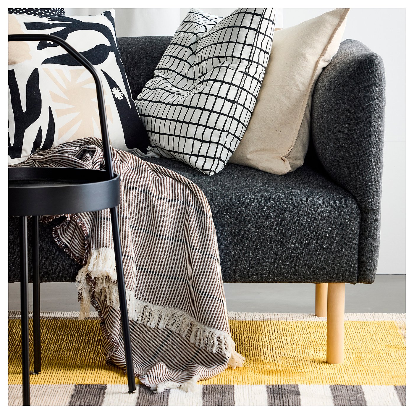 Modern grey sofa with black side table, patterned pillows, stripe blanket, and yellow plaid rug.