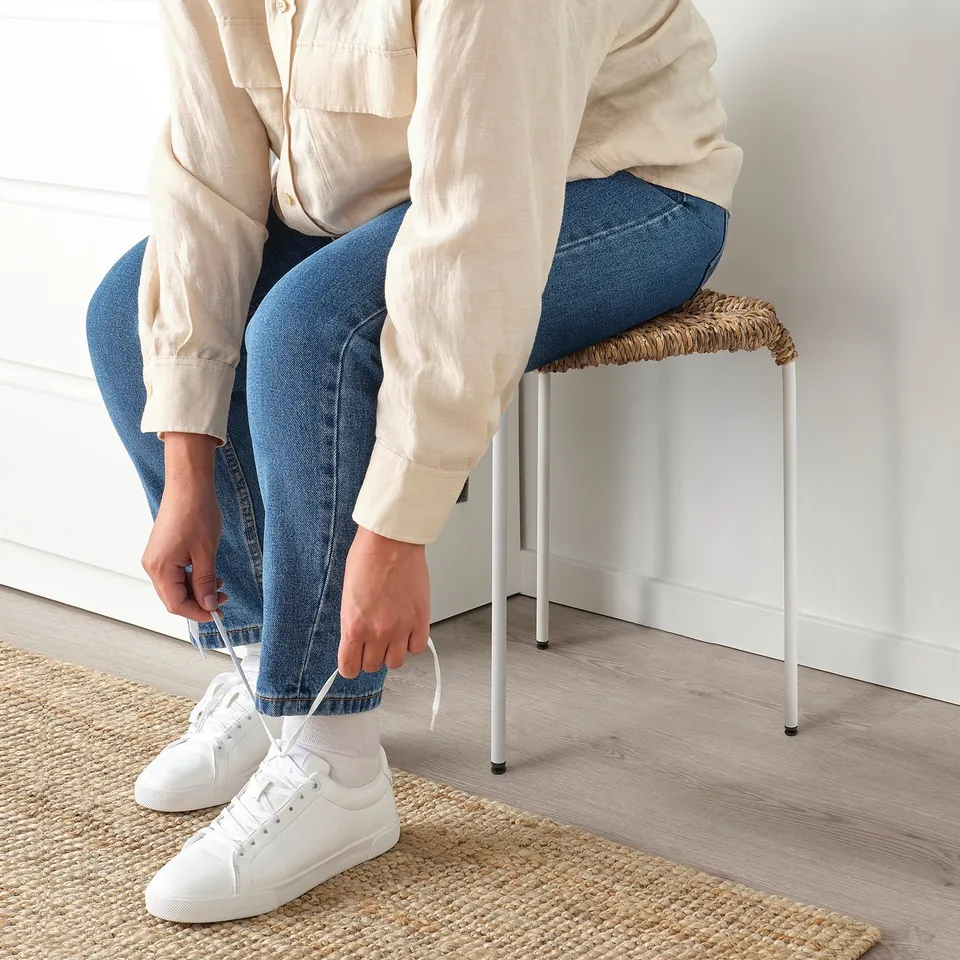 Person tying shoelace on a white trainer sitting on a woven, 3-legged stool.