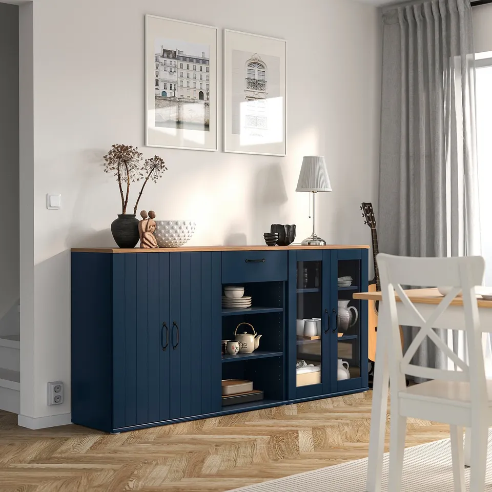 Modern dining area with blue SKRUVBY sideboard, featuring glass doors and wooden floors. White chair and lamp nearby, with wall art above.