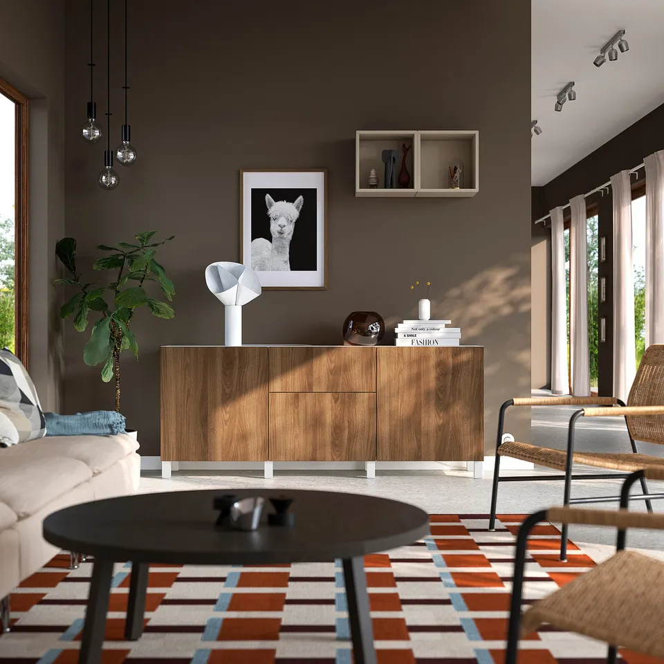 Modern living room with BESTÅ light brown sideboard, lamp, books, and decor. Wooden cabinetry, sofa, coffee table, large windows, and colourful rug.