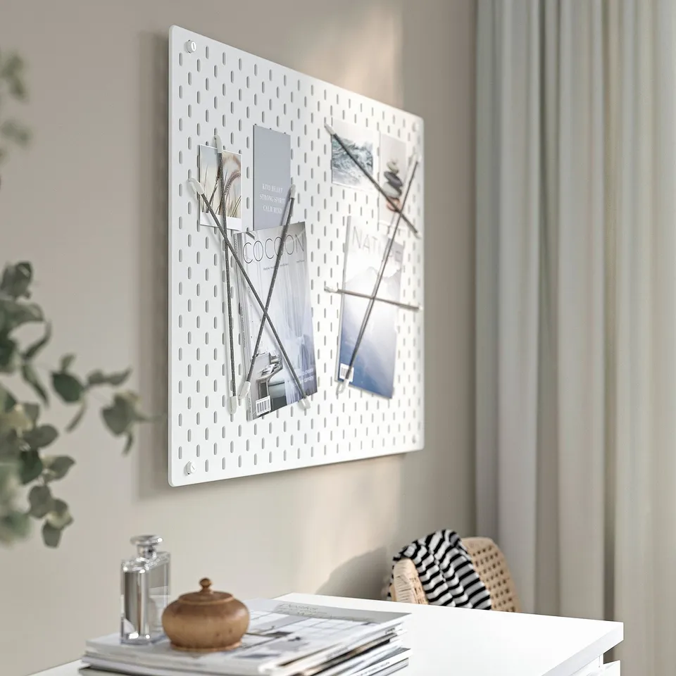 A white wall-mounted SKÅDIS pegboard displays organised photos and magazines. A stylish desk below holds a decorative plant, a jar, and a woven bag.