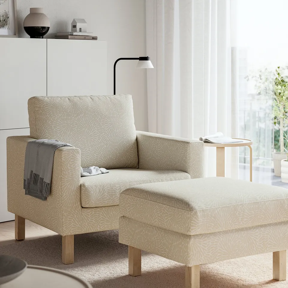 Beige patterned armchair & ottoman by window for relaxing.