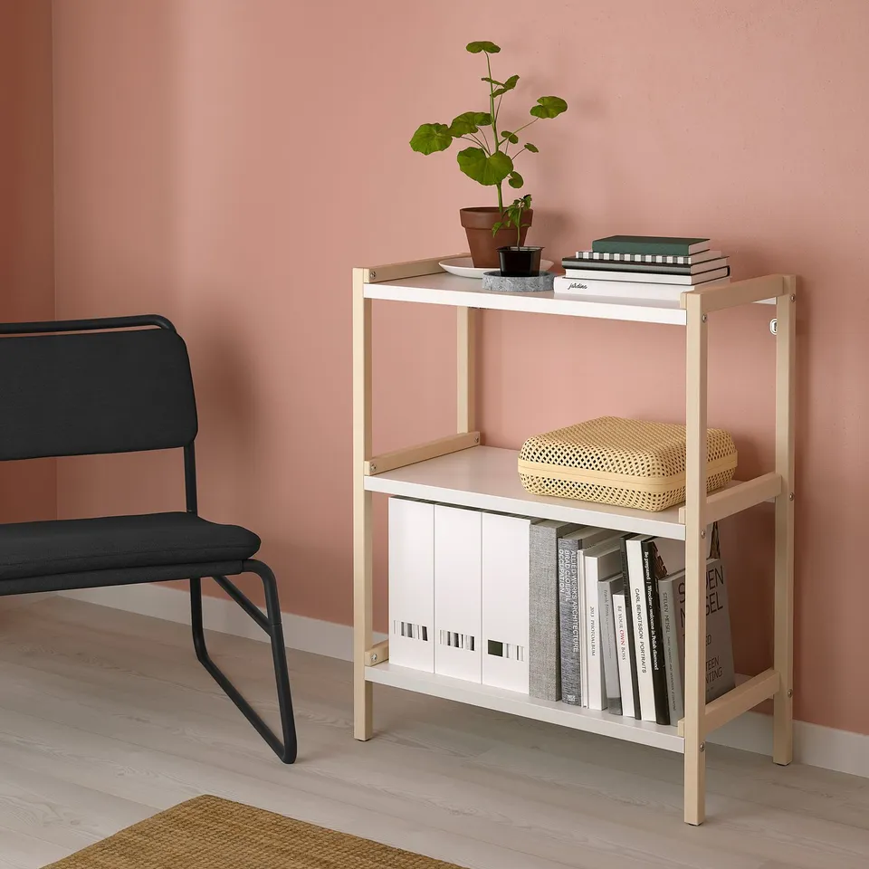 White 3-shelf EKENABBEN unit against pink wall holding books, plant, and basket; black chair beside.