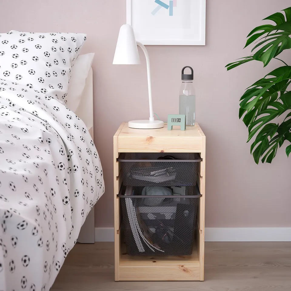Compact wooden TROFAST bedside table with mesh drawers, lamp, and water bottle, beside a bed with soccer-themed sheets.