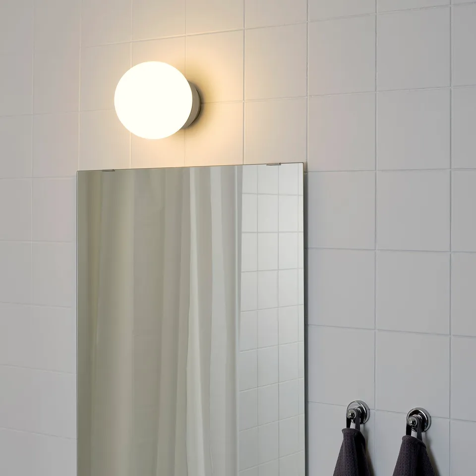 A round, wall-mounted lamp above a bathroom mirror, providing soft, general light. Black towels hang on metal hooks to the right.