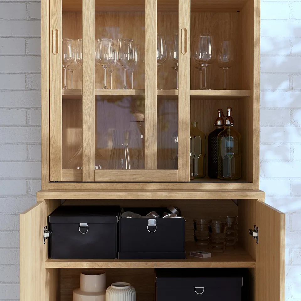 Wooden cabinet with glass doors, storing wine glasses, bottles, and boxes, displaying brush veneer and soft-close feature.