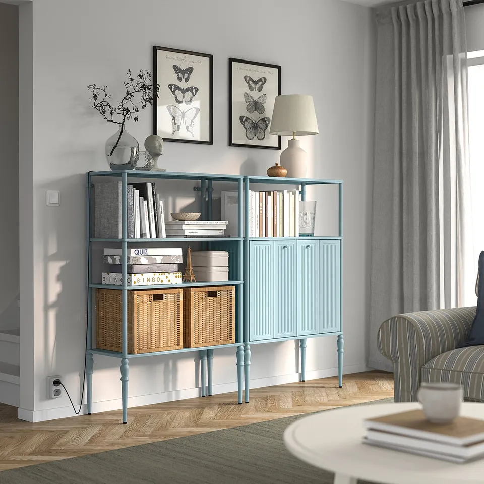 Light blue SÅGMÄSTARE metal cabinet and shelving unit with turned legs. Holds books and baskets, fits under large wall art, beside striped couch.