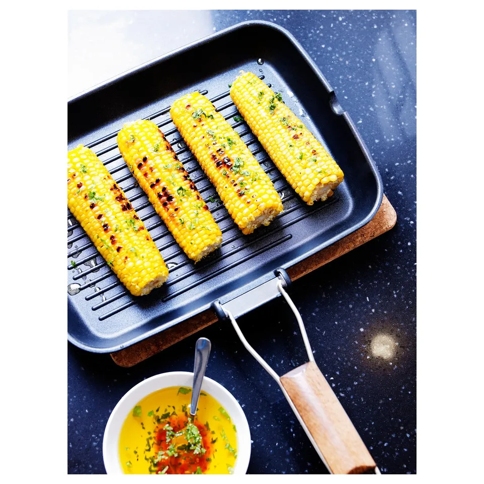 Four corn cobs grilling on a black GRILLA pan with a wooden handle and a yellow dipping sauce in a small bowl beside it.