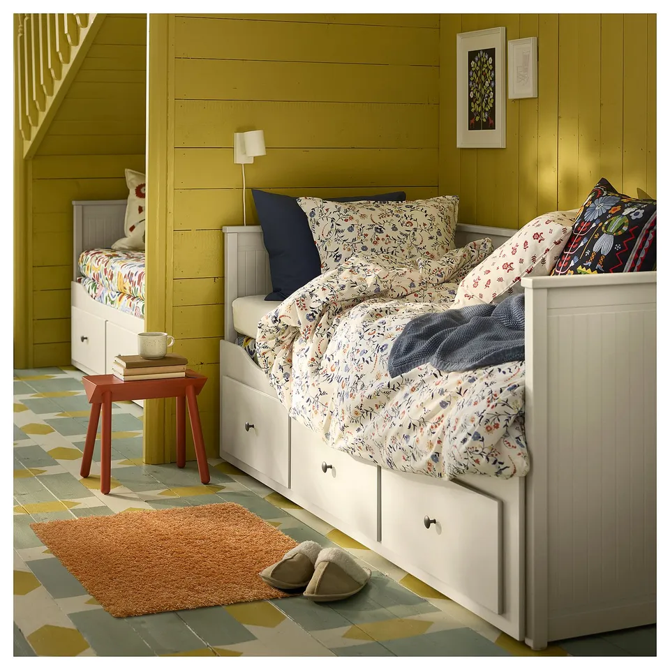 Cosy bedroom with white bed, floral linens, bedside table, lamp, rug, and slippers. Yellow walls, patterned floor.
