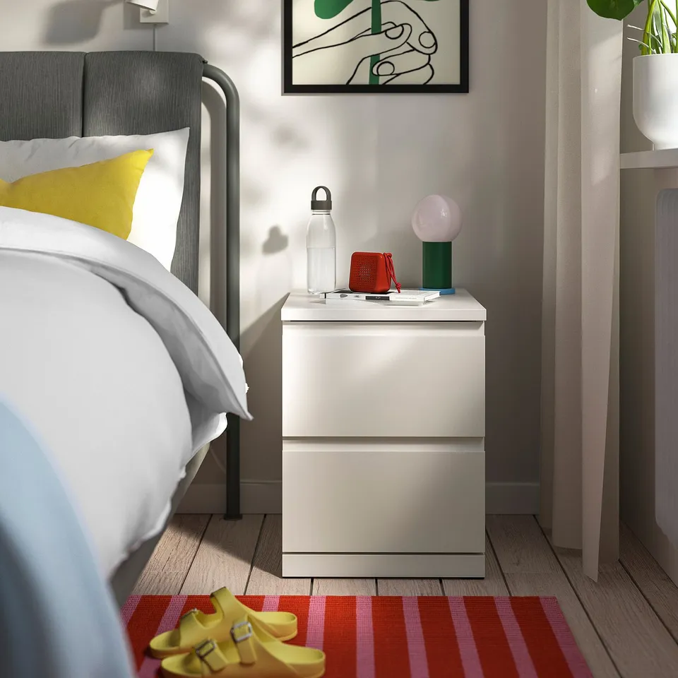 Modern bedroom with STORKLINTA bedside table. Drawer, bed with yellow pillow, water bottle, phone, wallet, lamp, red/pink rug, yellow sandals, plant.