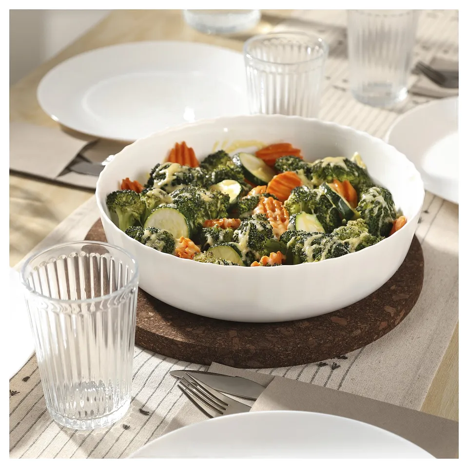 White oval ceramic dish filled with mixed vegetables on wooden table next to clear glasses and plates.