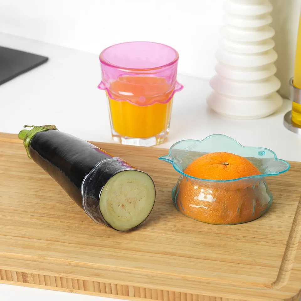 Wooden cutting board with sliced aubergine and an orange in a blue bowl, covered with pink and clear food savers.