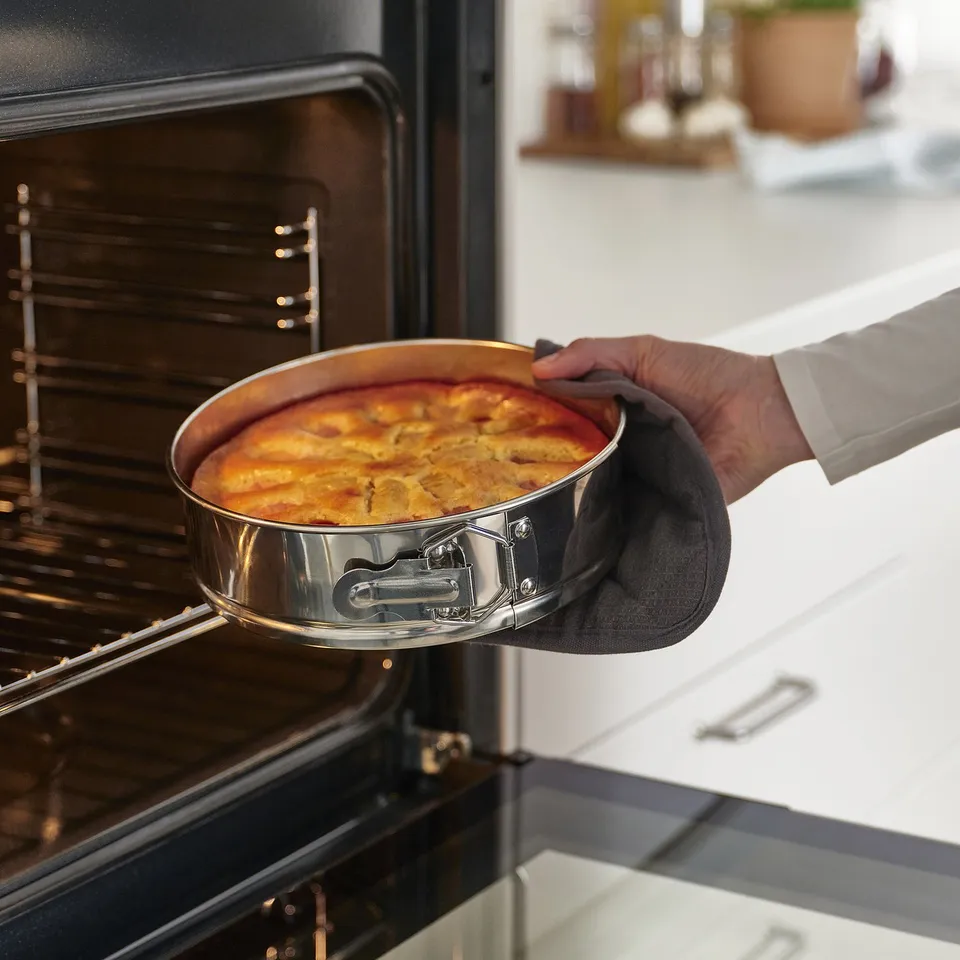 A hand holding a round springform pan with a baked item, removing it from an open oven. The pan a spring lock for easy release.