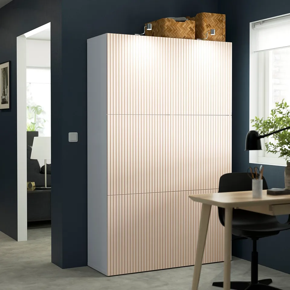 Modern office large light-colored BESTÅ storage unit with vertical lines, significantly taller than nearby desk and chair, topped with woven baskets.