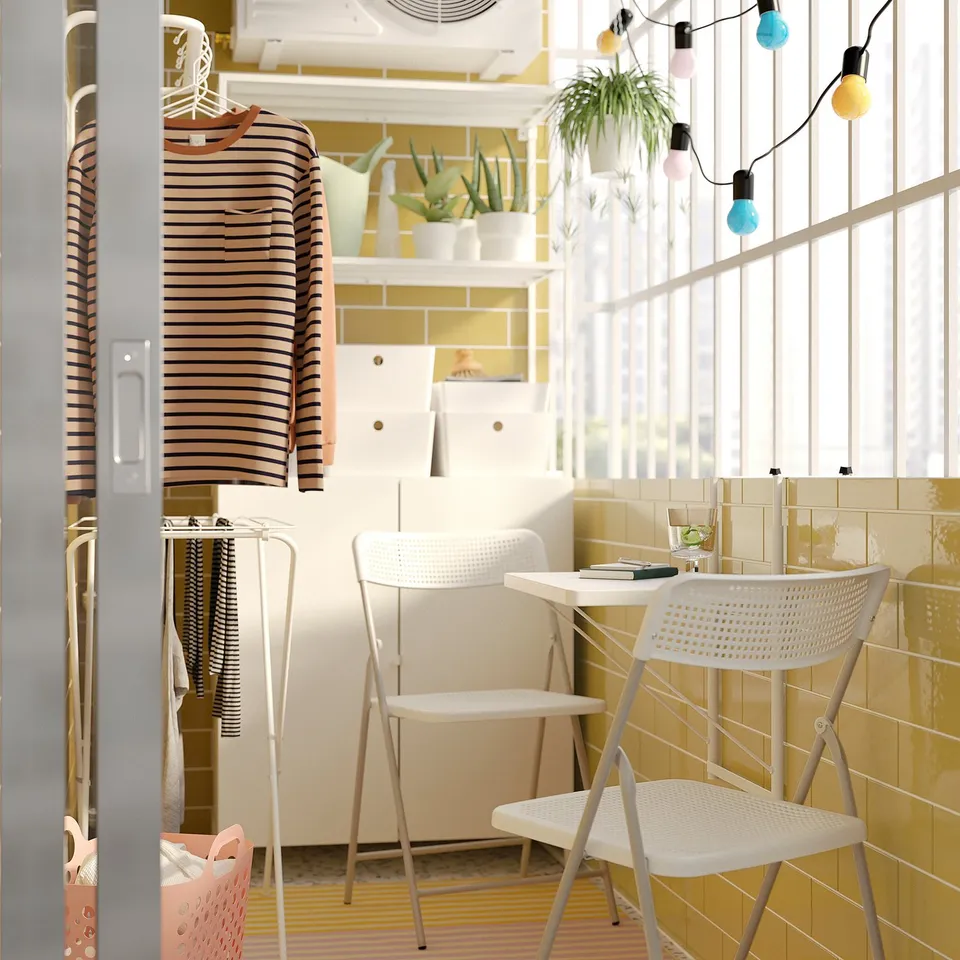 Bright balcony with white TORPARÖ chair and table. Colourful lights, plants, and striped shirt hanging.