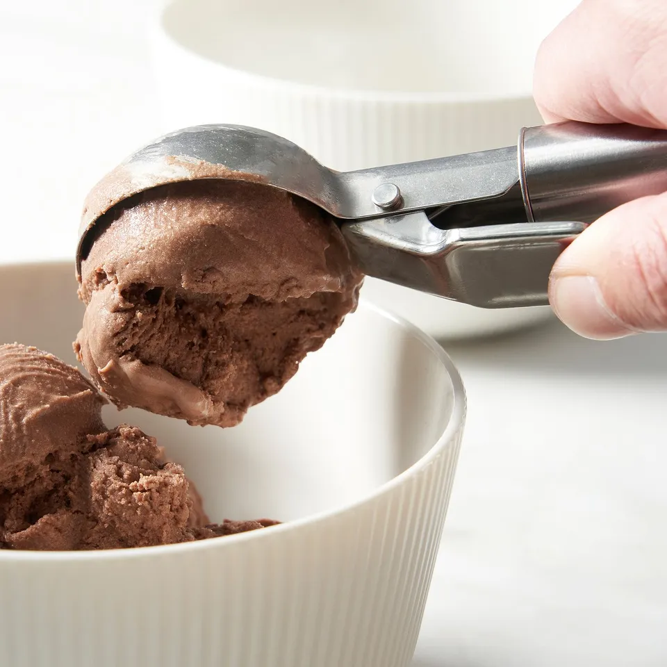 Hand scooping chocolate ice cream with a metallic tool into a white bowl. The tool has a lever for easy dispensing.