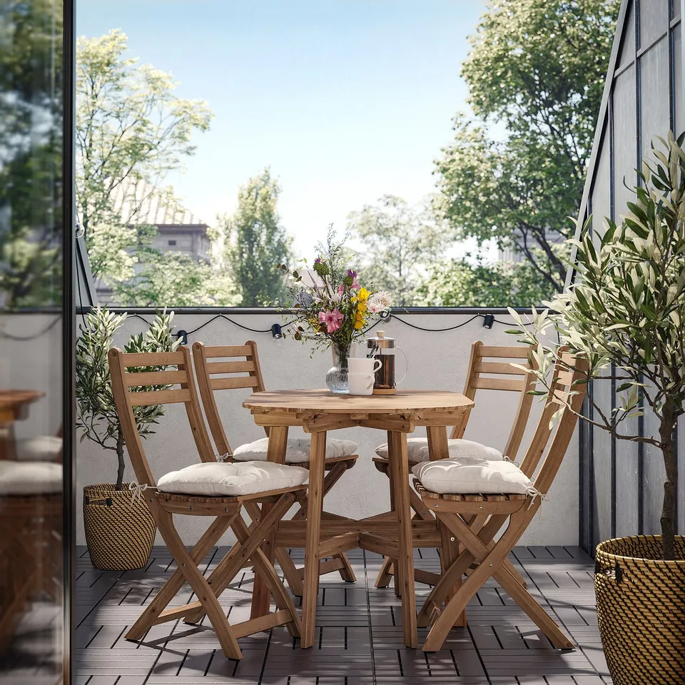 Small wooden outdoor table and chairs set with cushions on a balcony, featuring string lights and potted plants.