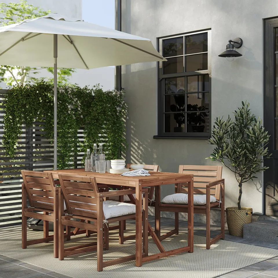 Backyard patio with NÄMMARÖ brown wooden table and four chairs. White cushions on chairs, an ivory umbrella, and a black house with a window.