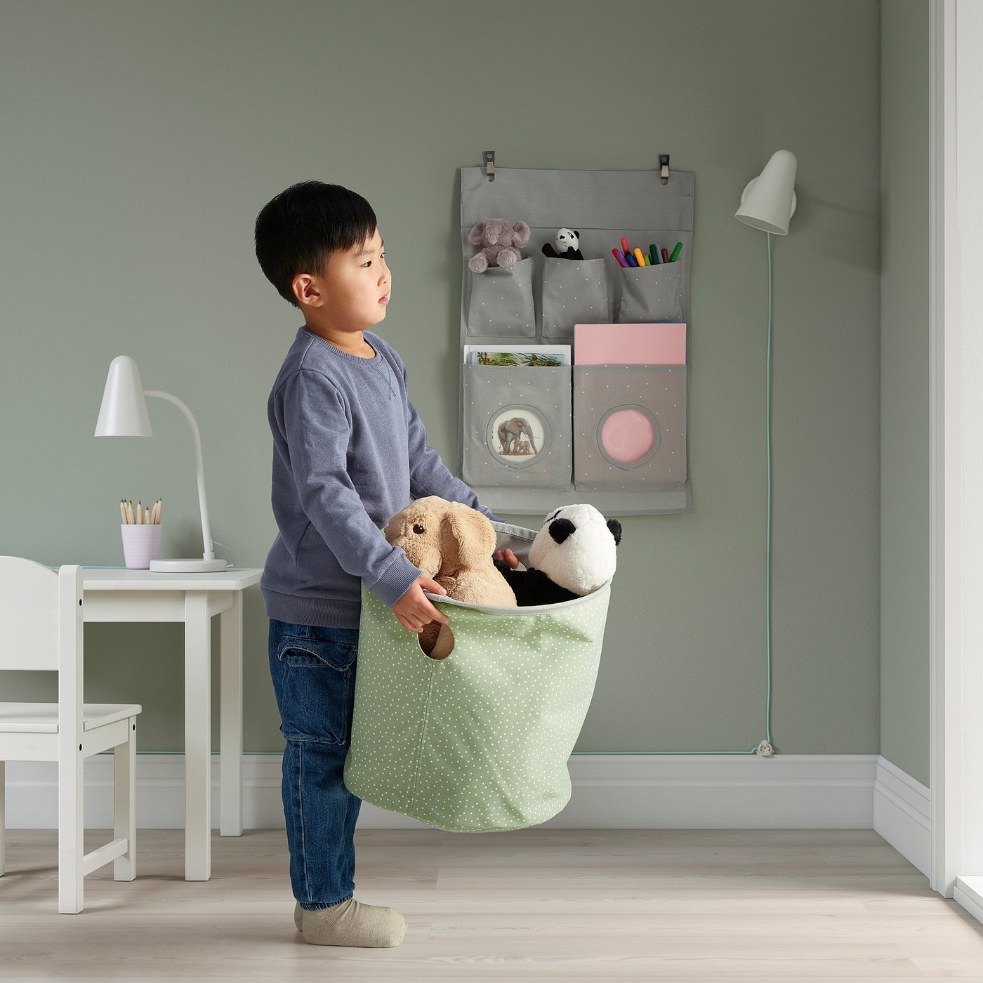 A child holding a green fabrics bag with stuffed animals. The wall behind has a hanging organiser with pockets and hooks visible.