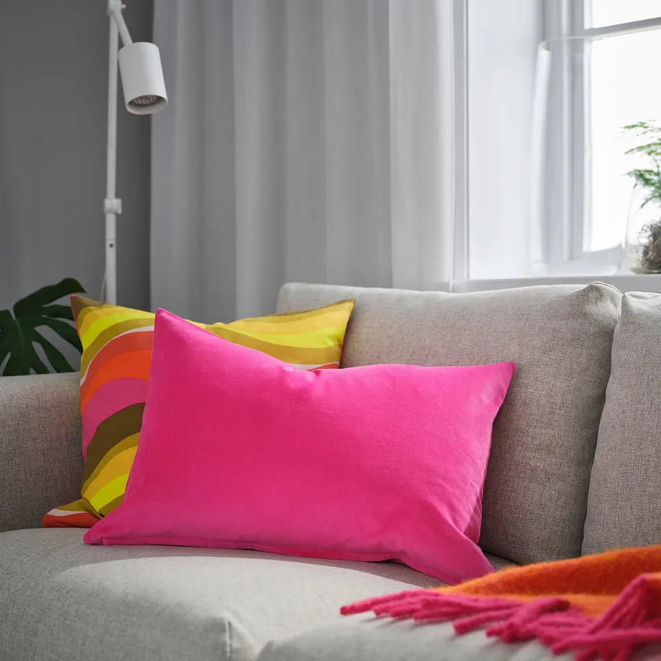 Bright pink SANELA cushion, large, soft with sheen, velvet texture. On a grey sofa, between a yellow pillow & near a window.