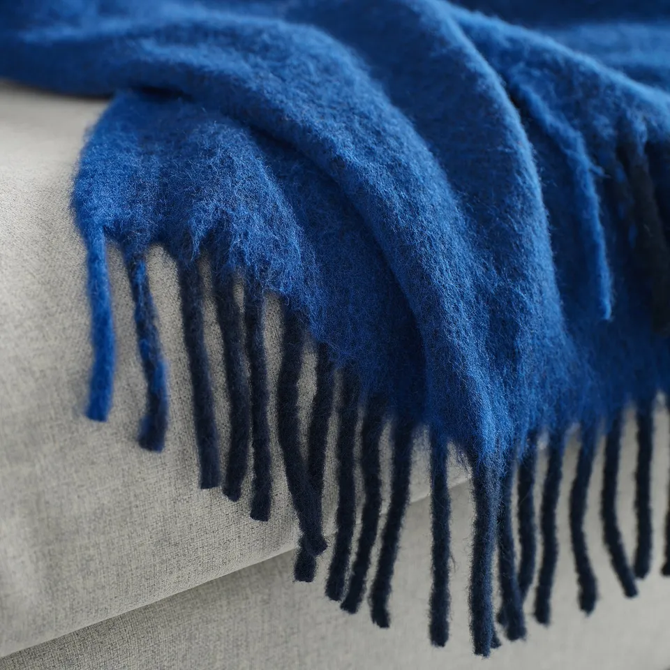 A close-up of a luxurious blue throw blanket with soft, fluffy texture and fringe ends, likely made of recycled polyester.