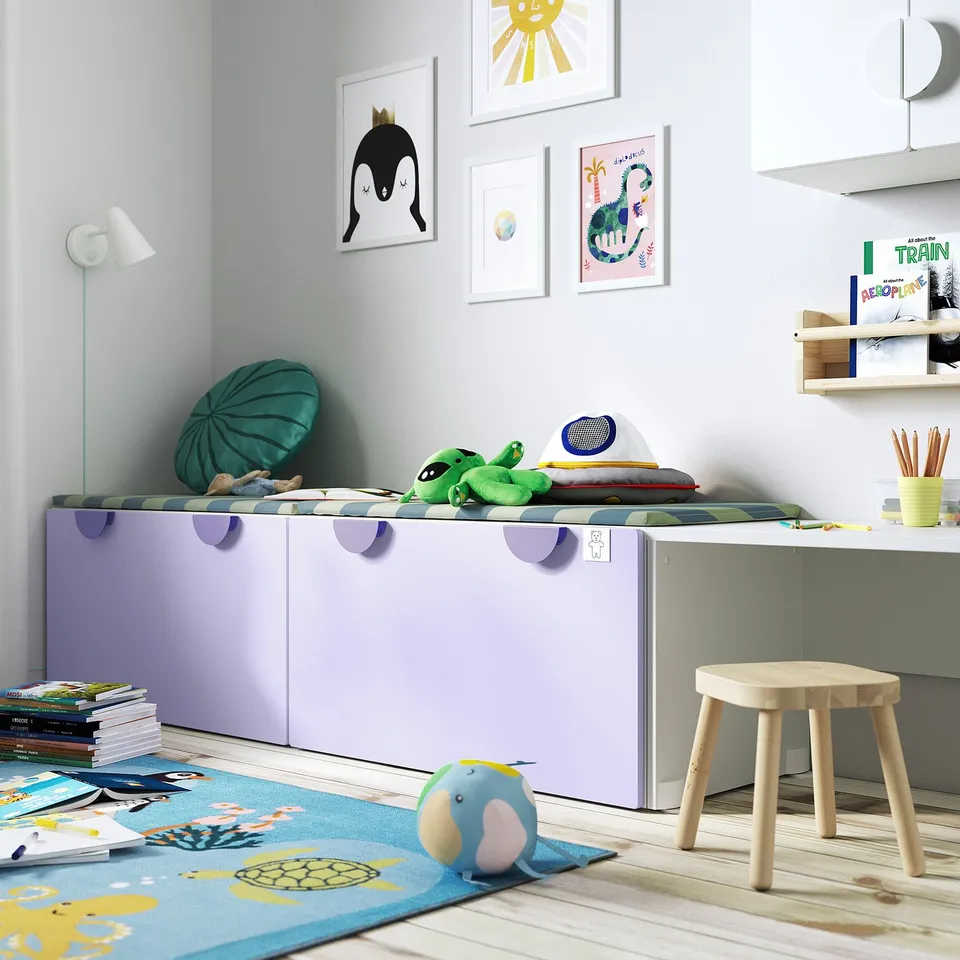 Childs white storage bench with cutouts, against wall with posters, between desk and lamp, with toys and blue rug.