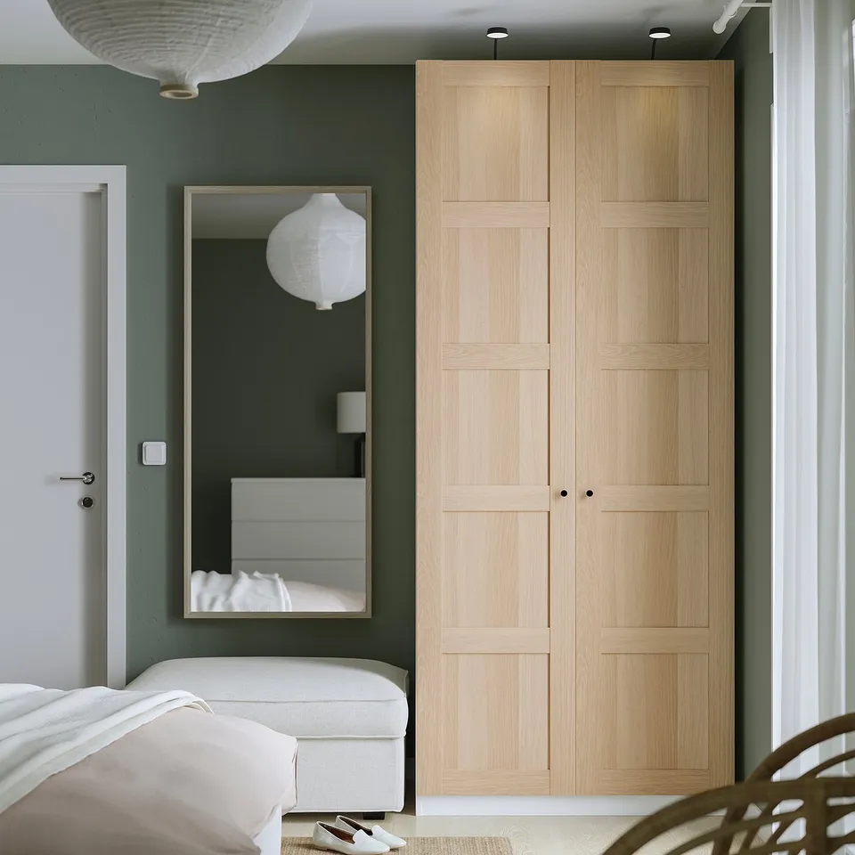 Beige PAX wardrobe with mirror, green wall. Tall, modern, sleek, wooden panels. Next to bed, taller than door.